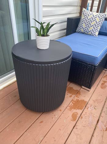 Ribbed outdoor side table with a plant on it, near a cushioned patio chair with a patterned pillow