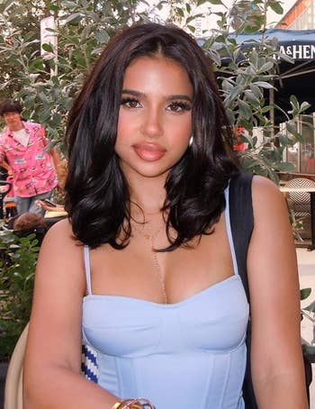 Person sitting outdoors in a city scene wearing a stylish outfit. Urban backdrop with trees and a cafe