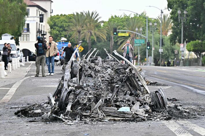Jornalistas relatam as cenas dos protestos em Los Angeles ao lado de um veículo queimado Foto: Frederic J. Brown/AFP