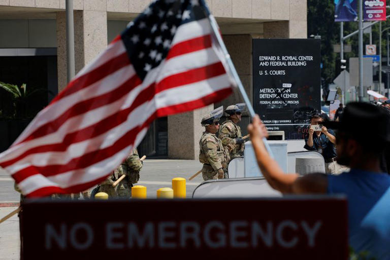 A man waves a U.S. flag as  of the California National Guard stand guard outside the Edward R. Roybal Federal building, after days of protests against federal immigration sweeps and the deployment of the California National Guard and U.S. Marines, in downtown Los Angeles, California, U.S., June 10, 2025.  REUTERS/Mike Blake