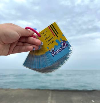 A hand holds a set of Poolside! card game cards with Taco Cat Goat Cheese Pizza text on the top card, near a beach backdrop