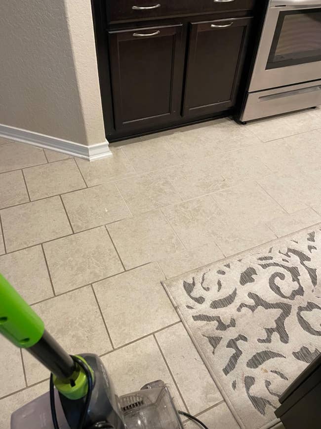 Vacuum cleaner on a tiled kitchen floor near cabinets and an oven. A patterned rug is partially visible