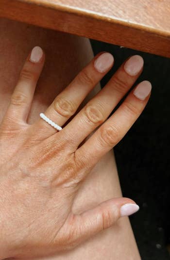 A hand with short, shiny nails wearing a simple beaded ring, resting on a person's leg, suggesting a minimalist fashion accessory style