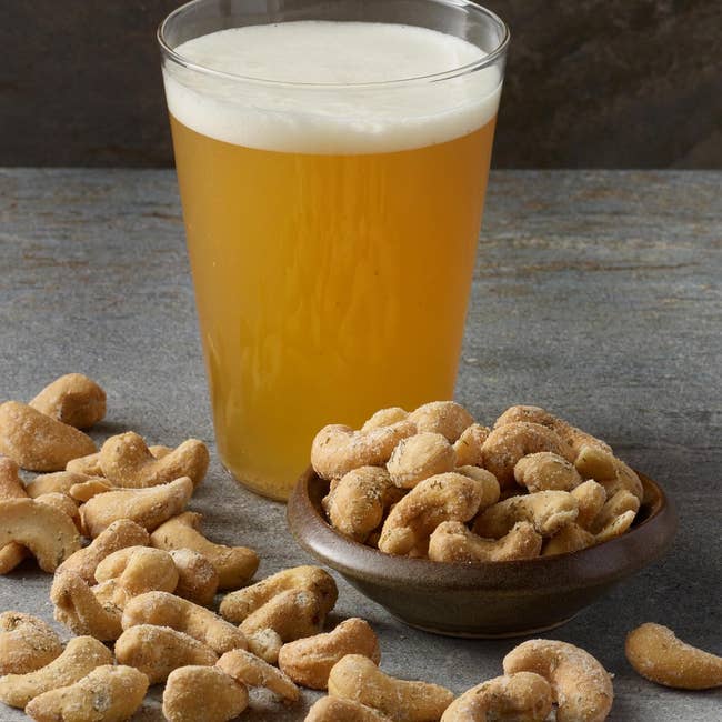 A glass of beer set next to a bowl and scattered cashews on a stone surface