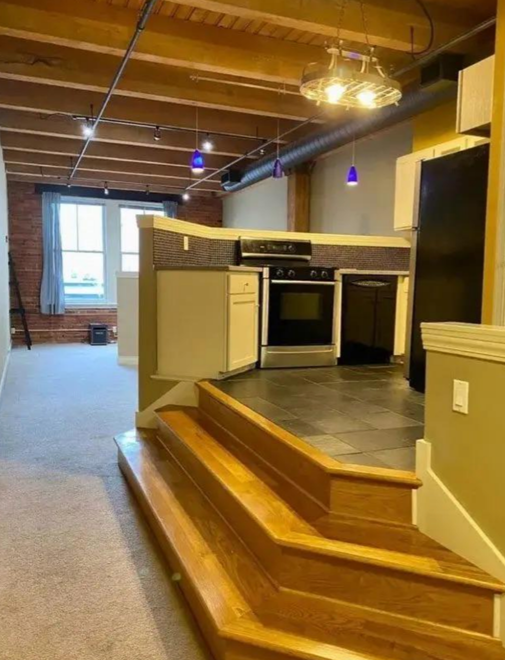 Kitchen slightly elevated with wooden steps leading up; features a stove, cabinets, and overhead lighting in a loft-style room with visible beams