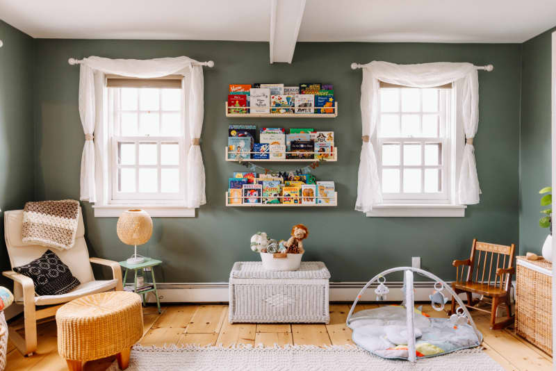 Children's room with green walls, bookshelves, wicker chair, play mat, and wooden rocking chair.
