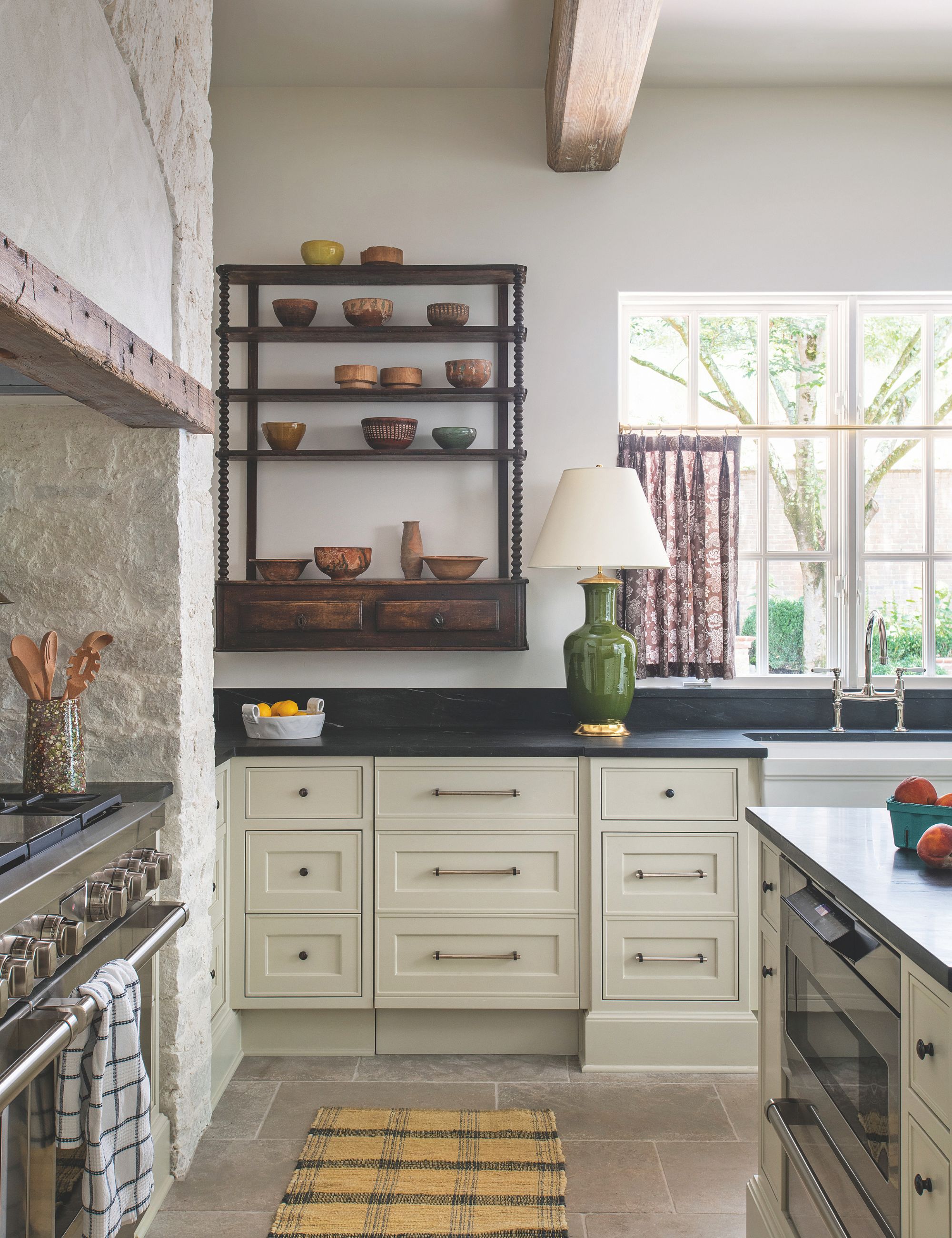 Cream kitchen with black worktops a wooden shelving unit on the wall and a large stone fireplace