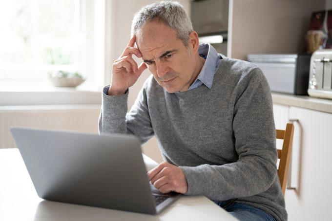 A person focuses on a laptop screen, sitting at a kitchen table, with a thoughtful expression.