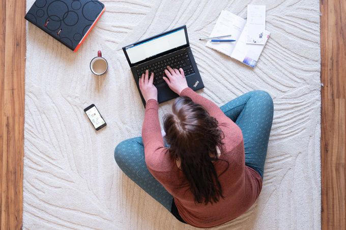 Person typing on a laptop seated on a patterned rug, surrounded by a phone, notebook, pen, cup, and binder, in a wooden-floored room.