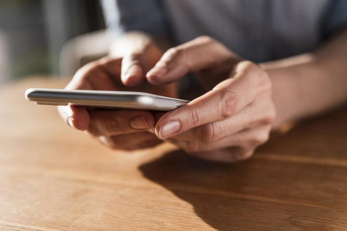 Hands hold a smartphone, tapping the screen while seated at a wooden table.