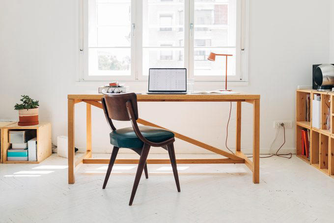 A laptop sits on a wooden desk near a window, accompanied by a chair, books, a lamp, and a potted plant in a minimal room.