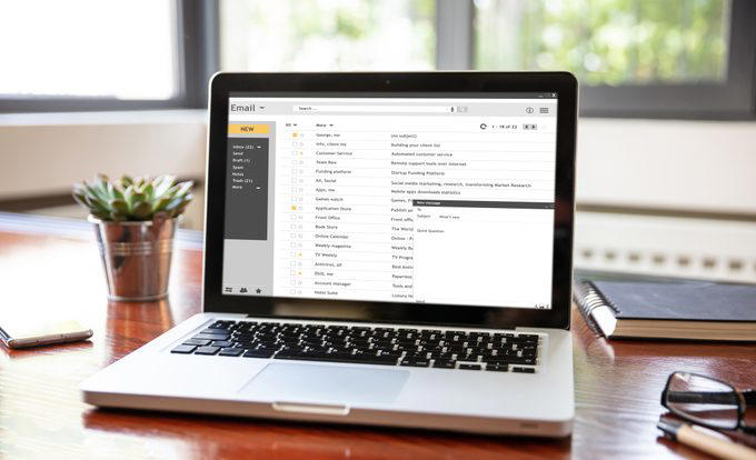 Laptop displays an email inbox with various messages in a bright office, next to a plant, notebook, glasses, and smartphone on the desk.