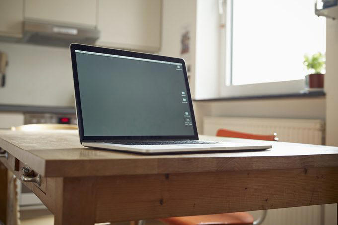 Laptop sits open on a wooden table in a modern kitchen, with natural light streaming through a window and a plant nearby.