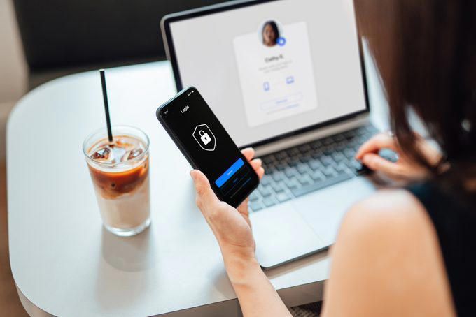 Phone displays login screen; person types on a laptop. A glass of iced coffee with a straw sits nearby on the table.