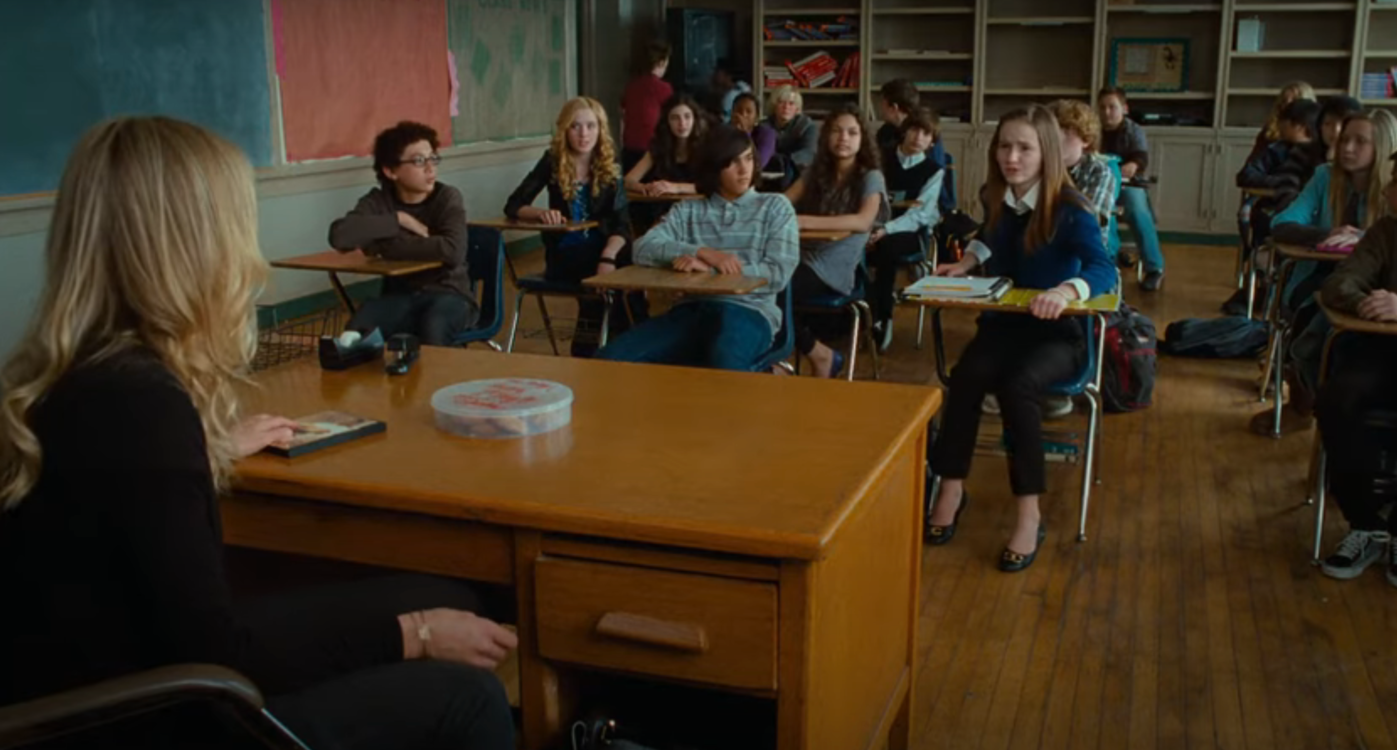 Classroom scene with students seated at desks facing a teacher at a wooden desk, illustrating a typical school setting