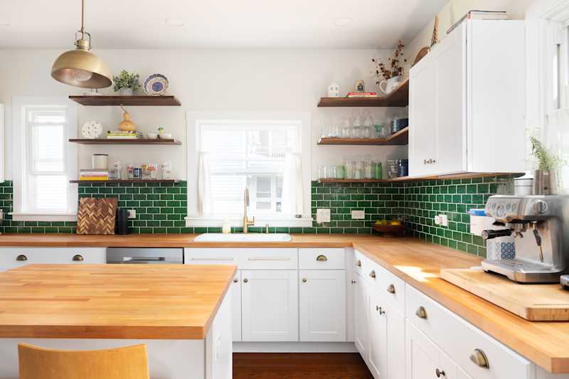 ELMHURST, IL, USA - MAY 25, 2021: A renovated kitchen with white cabinets, a natural wood countertop, chairs sitting at the island, a green subway tile backsplash, and a gold pendant light.