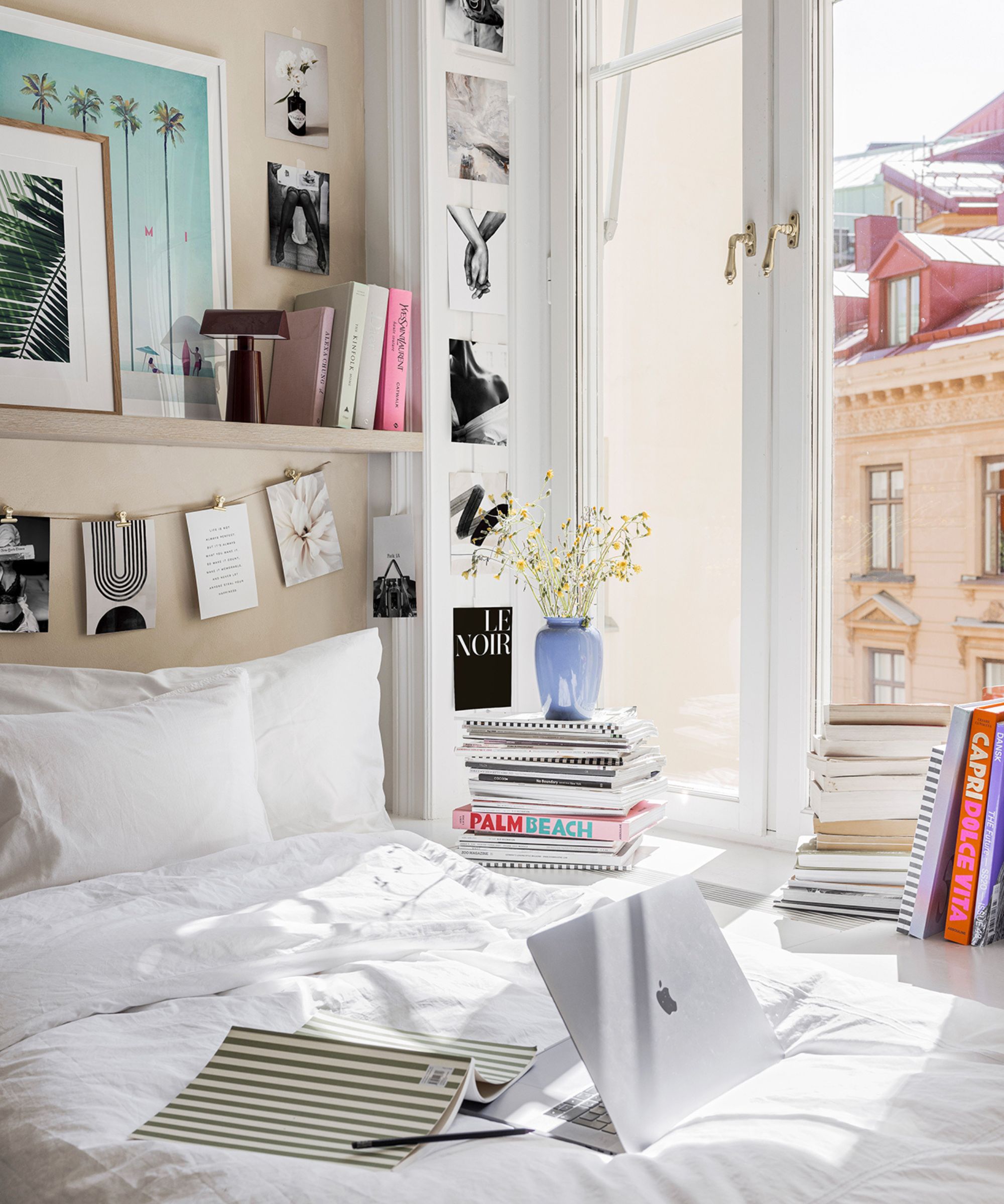 A corner dorm room bed against a large window. A Macbook on the bed next to an open notebook. black and white photo prints on the wall and hanging from a peg line. A shelf above with books and photographs.