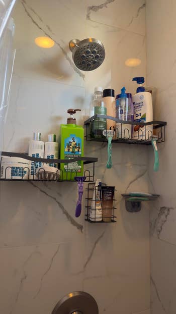 Shower area with marble walls, two shelves holding various bottles of shampoo, conditioner, body wash, and razors, under a chrome showerhead