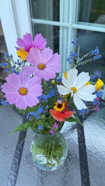 Vase with assorted wildflowers on a glass table, placed in front of a window; suitable for home decoration shopping article