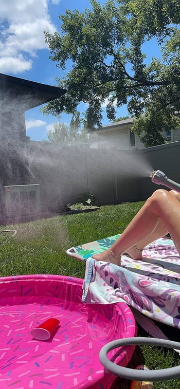 Person lounging on a towel in a backyard, spraying water mist, with a pink kiddie pool and houses visible. Ideal setting for outdoor relaxation products