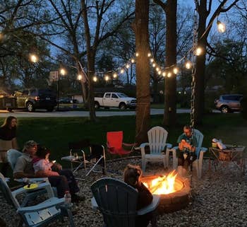 Family seated around a fire pit in a backyard at dusk, under string lights, with trees and parked vehicles in the background