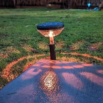 A garden solar light illuminates a small area of grass and concrete at dusk, casting a soft shadow