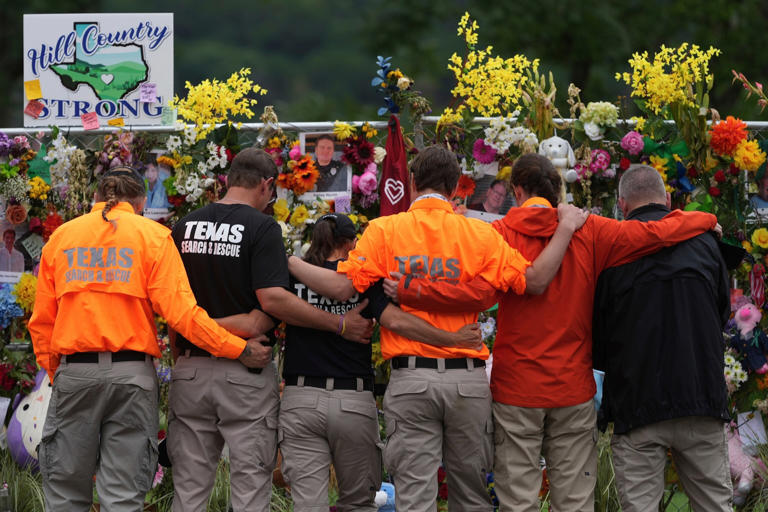 Eric Gay/AP - PHOTO: Members of a search and rescue team embrace as they visit a memorial wall for flood victims, July 13, 2025, in Kerrville, Texas.