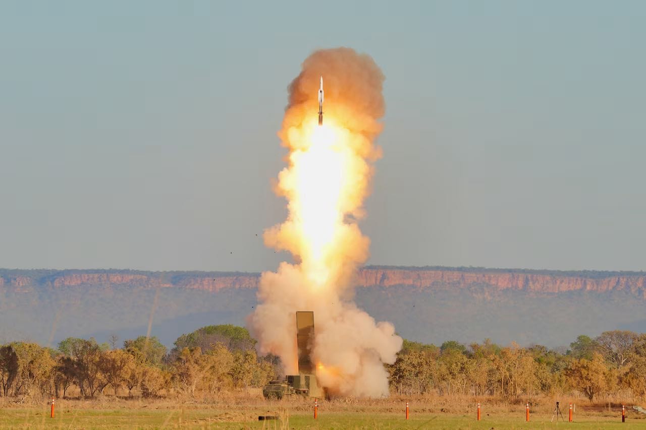 A missile is shot vertically from a launcher. The ground is grassy terrain with some trees. In the background is a line of mountainous formations and a hazy blue sky.