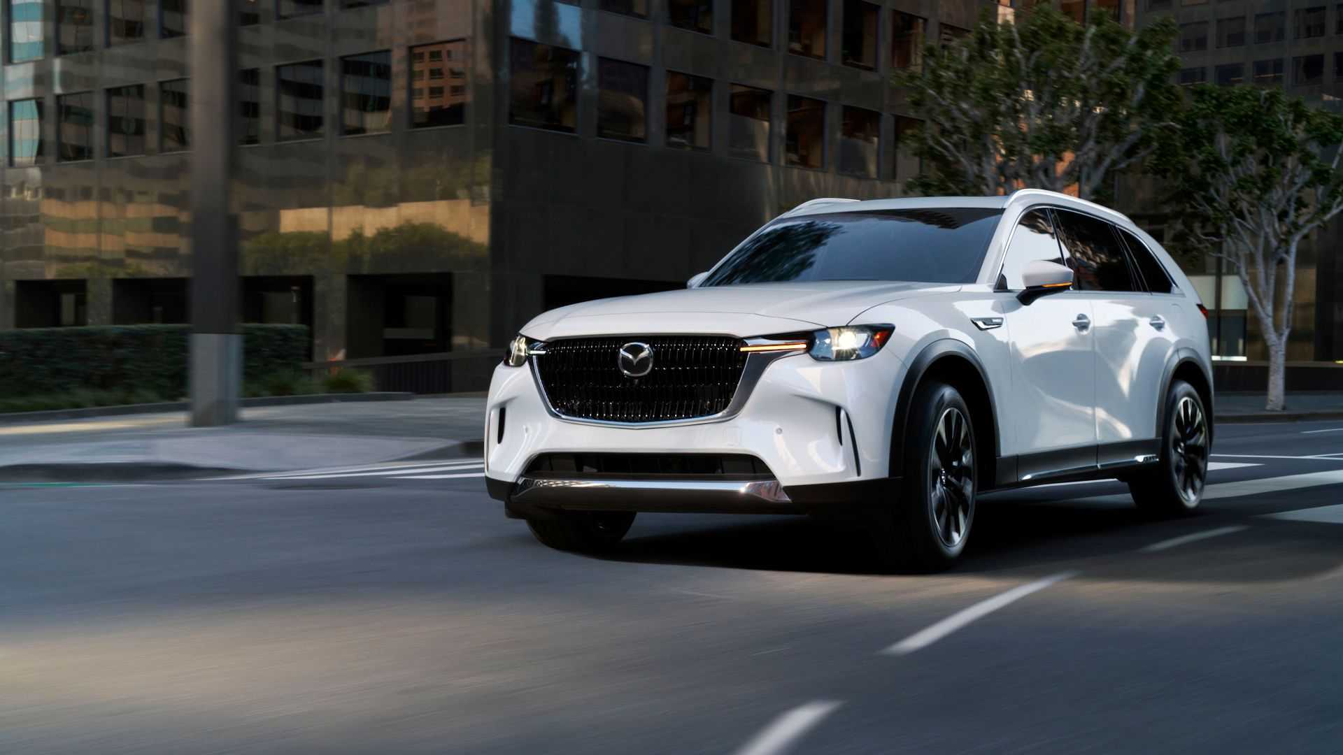 Dynamic front 3/4 shot of a white 2025 Mazda CX-90 driving in a city.