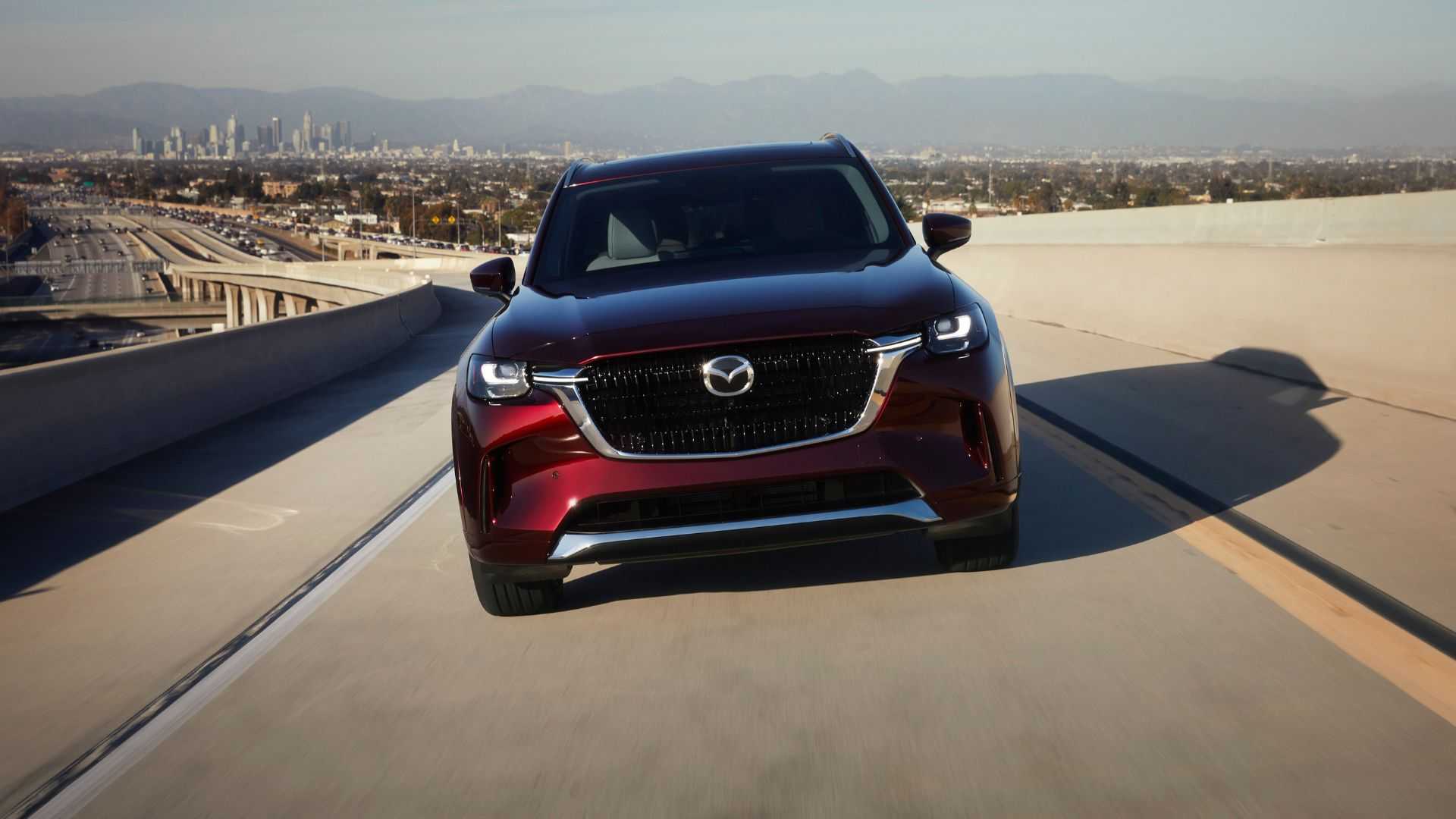 Dynamic front-end shot of a red 2025 Mazda CX-90 driving on a freeway with a cityscape in the background.