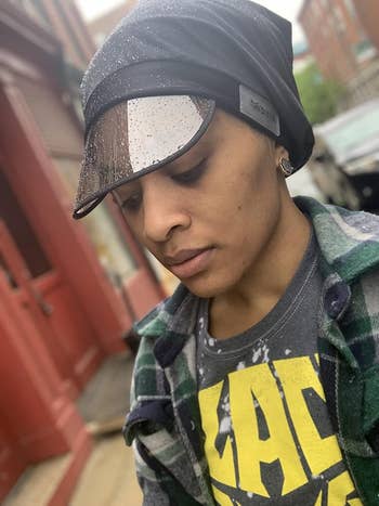 Person wearing a rain visor cap and plaid jacket stands outside a storefront. Text on shirt partially visible. Shopping article context