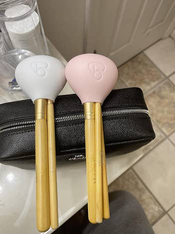 Two cosmetic brushes with light and pink bulbous tops, wooden handles, on a black toiletry bag. On a bathroom counter