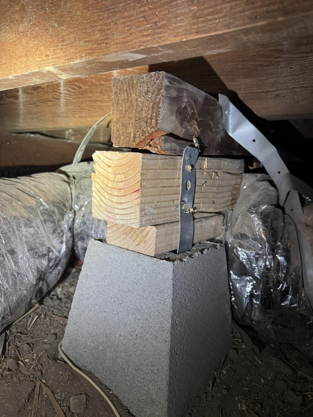 Wooden blocks and a metal strap support a concrete pier under a house; plastic-wrapped insulation is visible around them