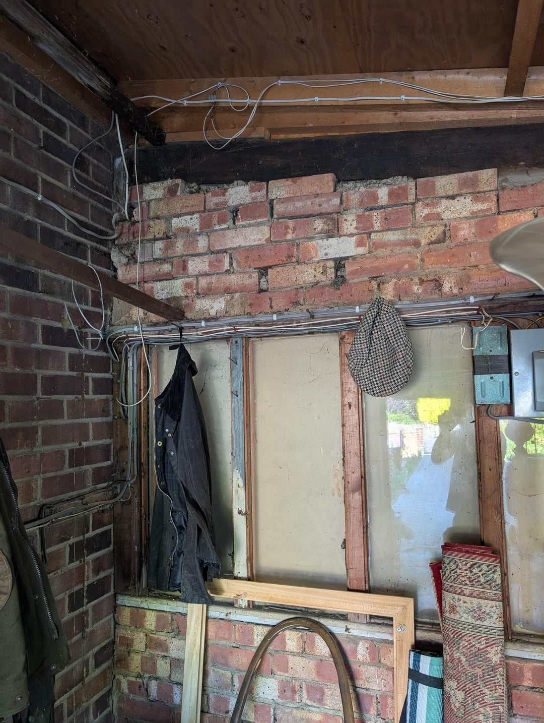 A cluttered garage corner with hanging jackets, a hat, exposed brick walls, cables, and various stored items