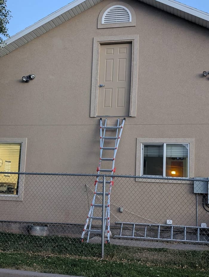 A ladder propped on a fence leads to a door on the second story of a house with no balcony or visible platform