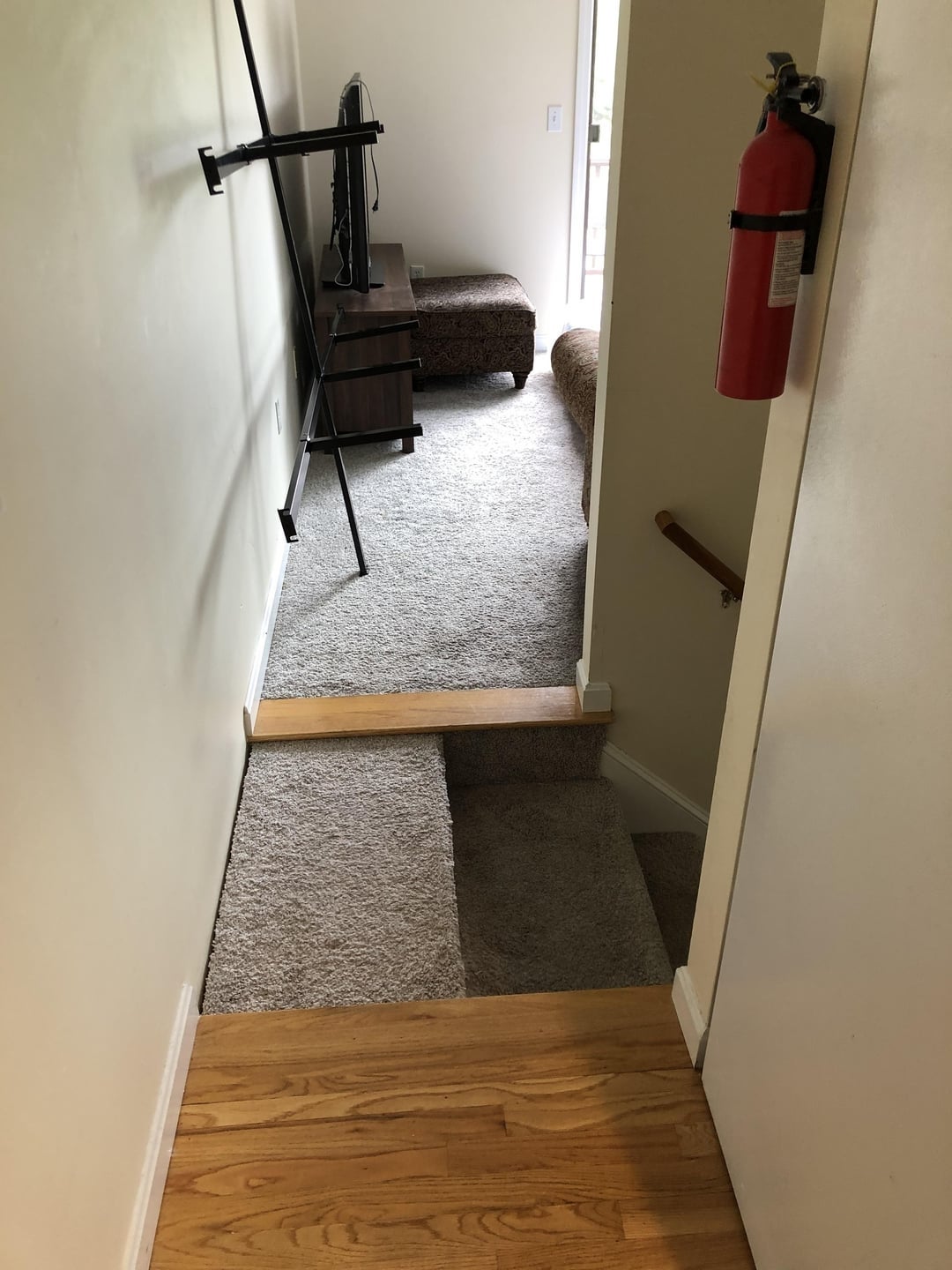 Stairs with uneven step height leading to a carpeted room, featuring a mounted TV and a fire extinguisher on the wall