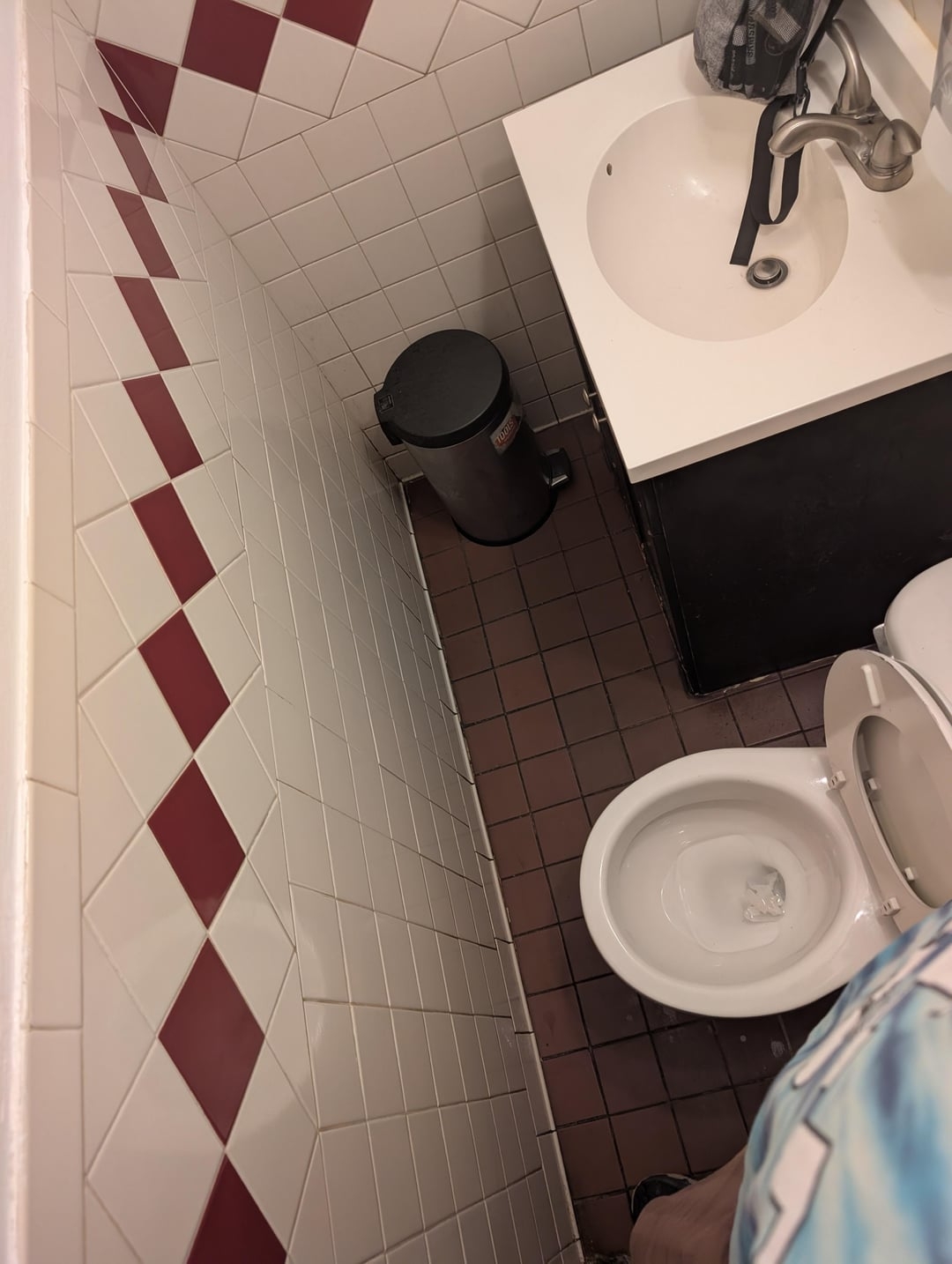 Bathroom with limited space, showing a toilet, sink, and trash can. The small size makes movement challenging