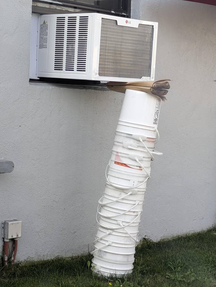 An air conditioner is propped up by a stack of white buckets, creatively supported against a wall