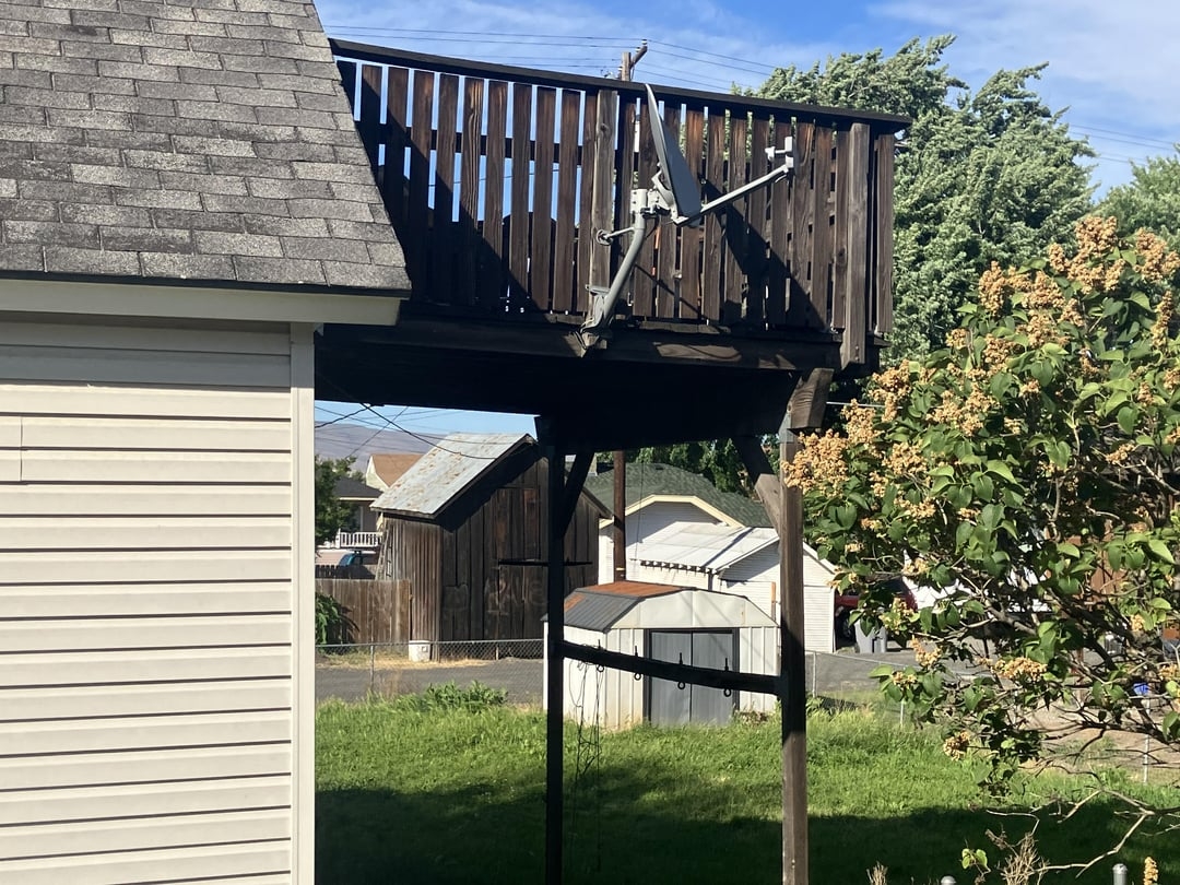 A rooftop deck with a satellite dish is built atop wooden supports, extending from a house. A garden and shed are visible in the background