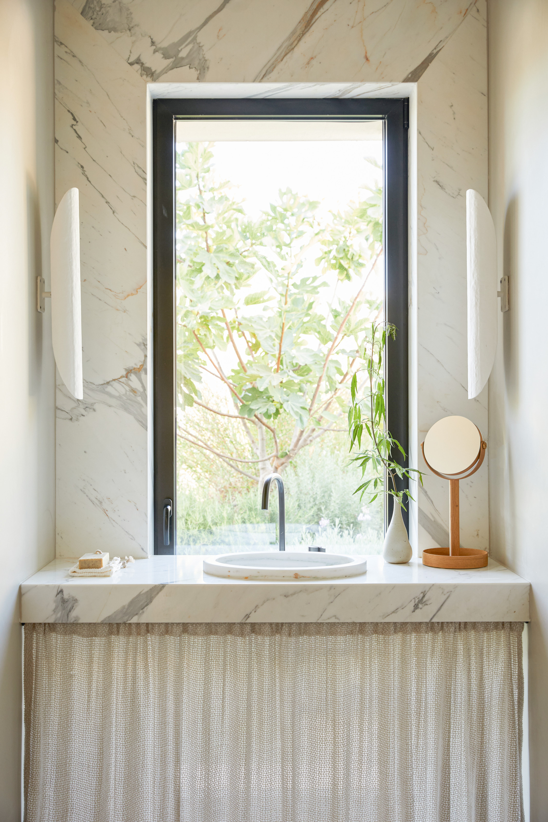 Bathroom vanity with white marble surfaces, slimline wall lights, inset round marble sink and beige linen cupboard curtain