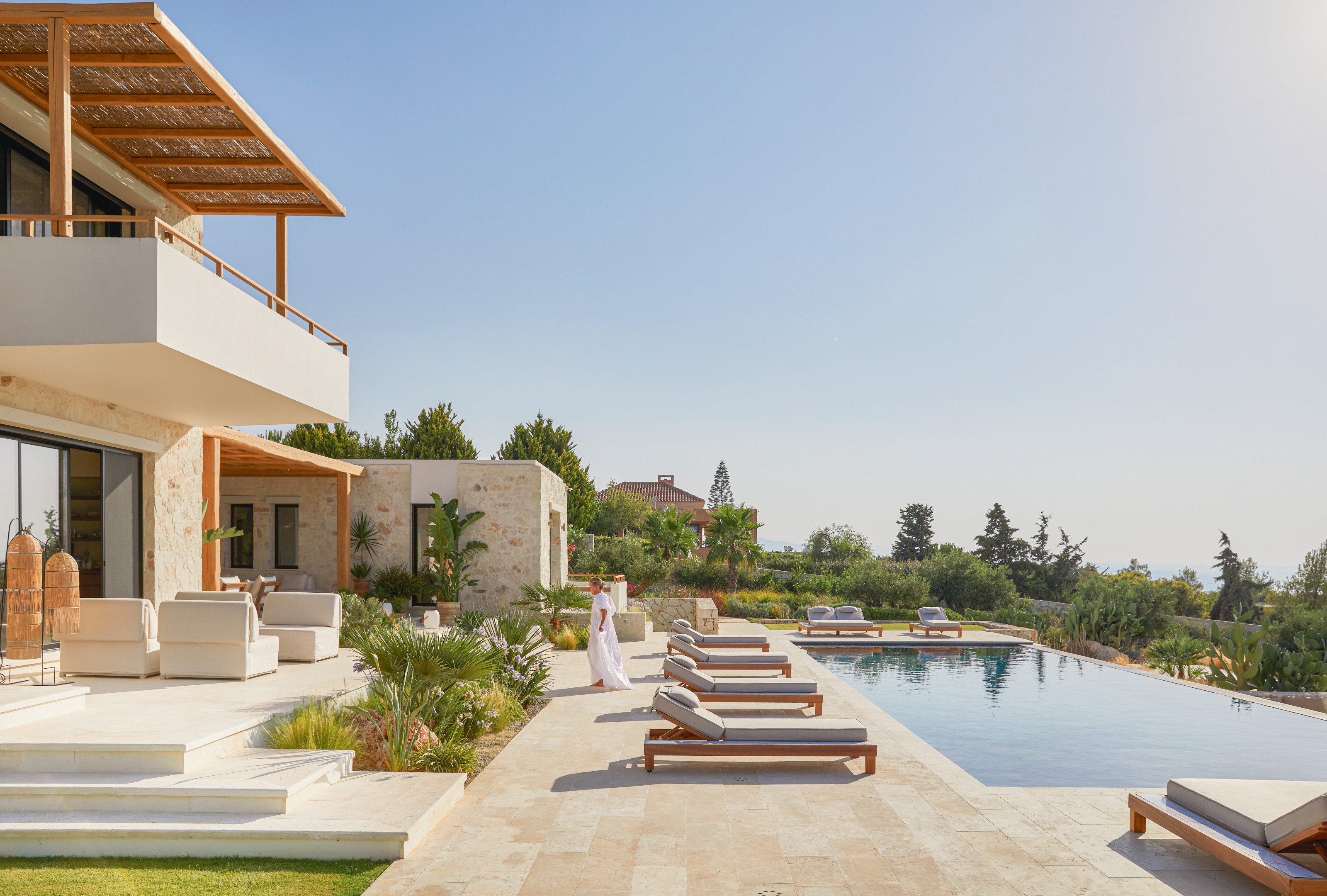Pool area of the garden with an infinity pool, beige stone paving, sun loungers and white outdoor armchairs