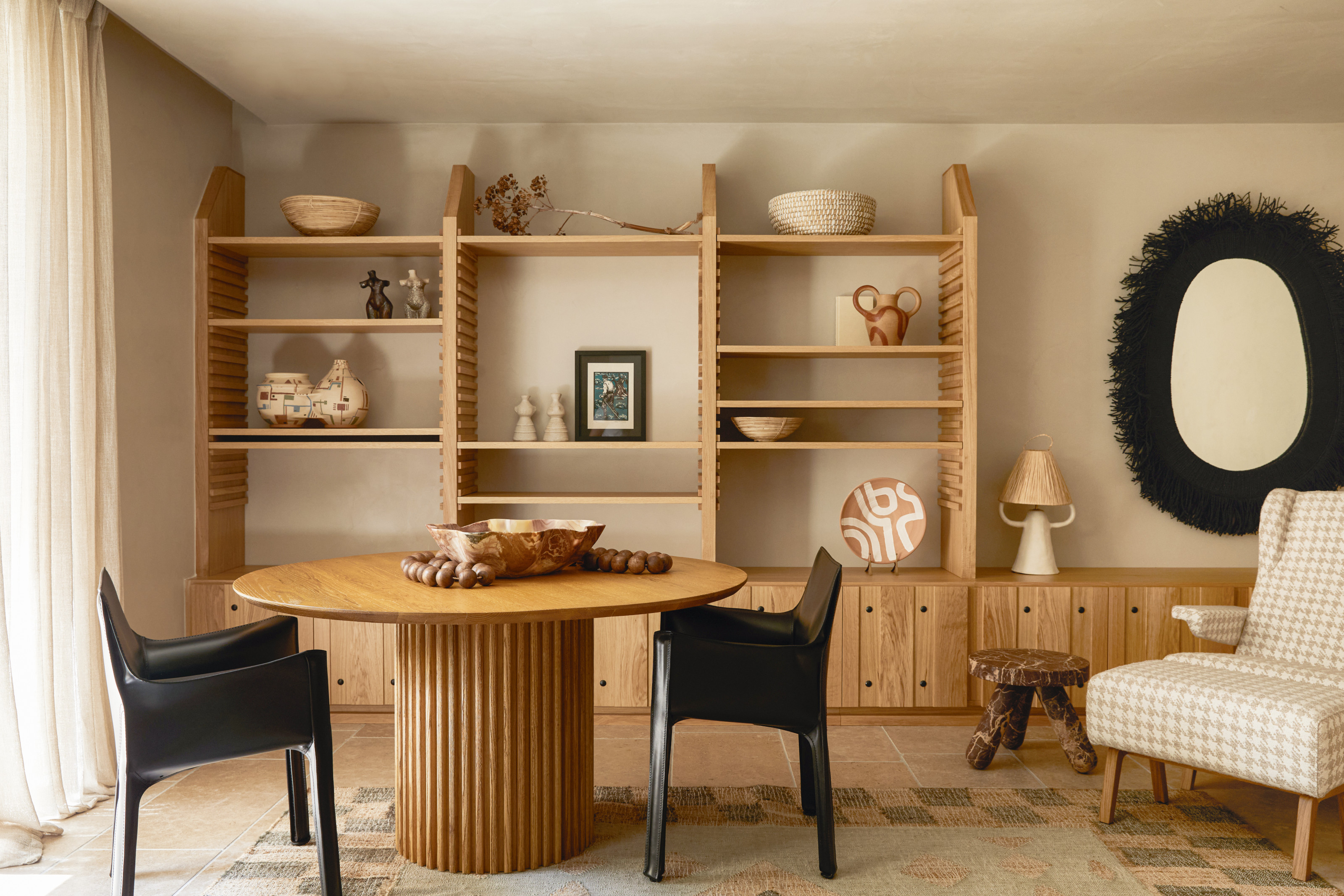 Dining space with wooden open shelving system, wood pedestal table, black leather dining chairs and a beige gingham armchair