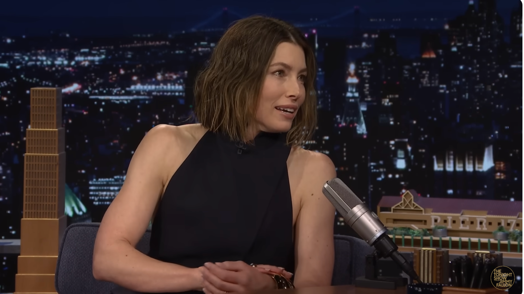 Jessica Biel with shoulder-length hair in a sleeveless, high-neck dress sits on a talk show set at night, speaking into a microphone
