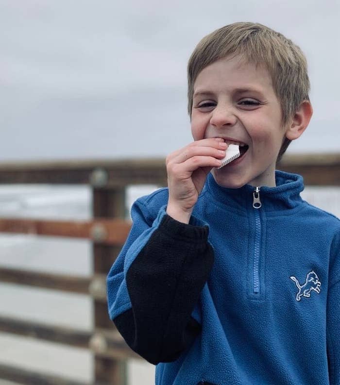 A smiling child in a blue fleece jacket enjoys a snack on a wooden pier by the sea