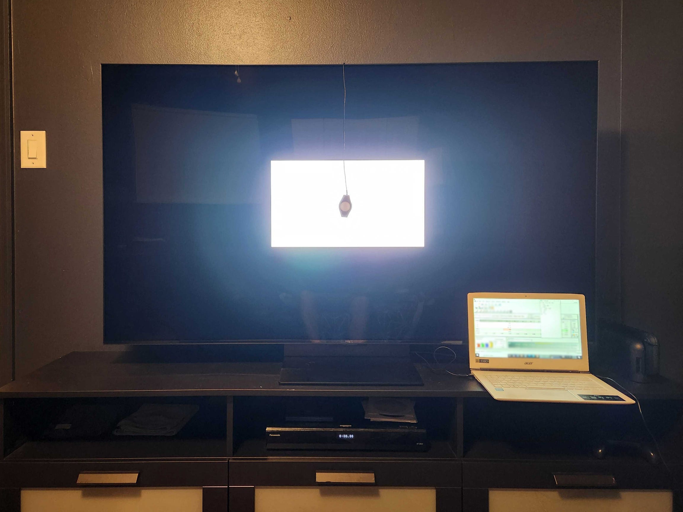 A TCL QM7K TV on a media console with a colorimeter placed on a white box displayed on the screen.