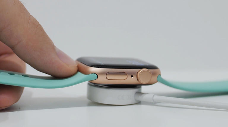 A hand placing an Apple Watch with a teal band onto a magnetic charging puck