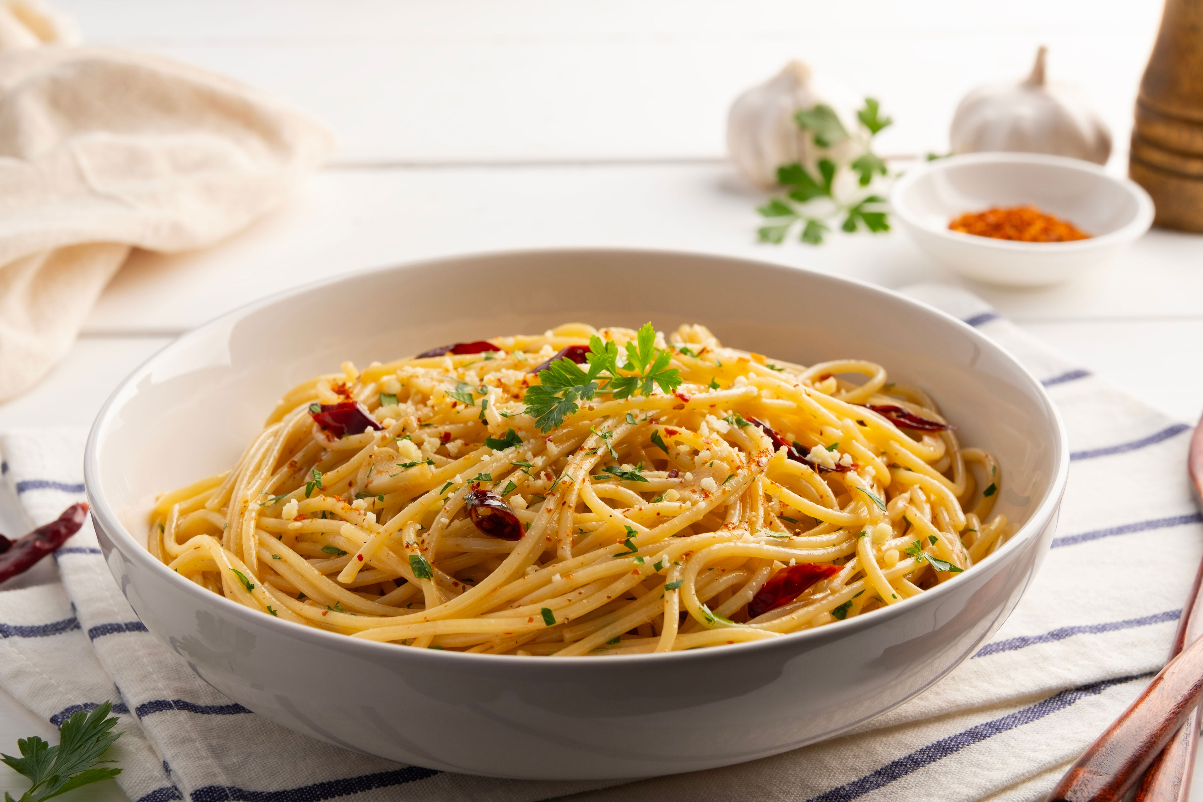 A bowl of spaghetti aglio e olio garnished with parsley, set on a table with garlic and spices in the background