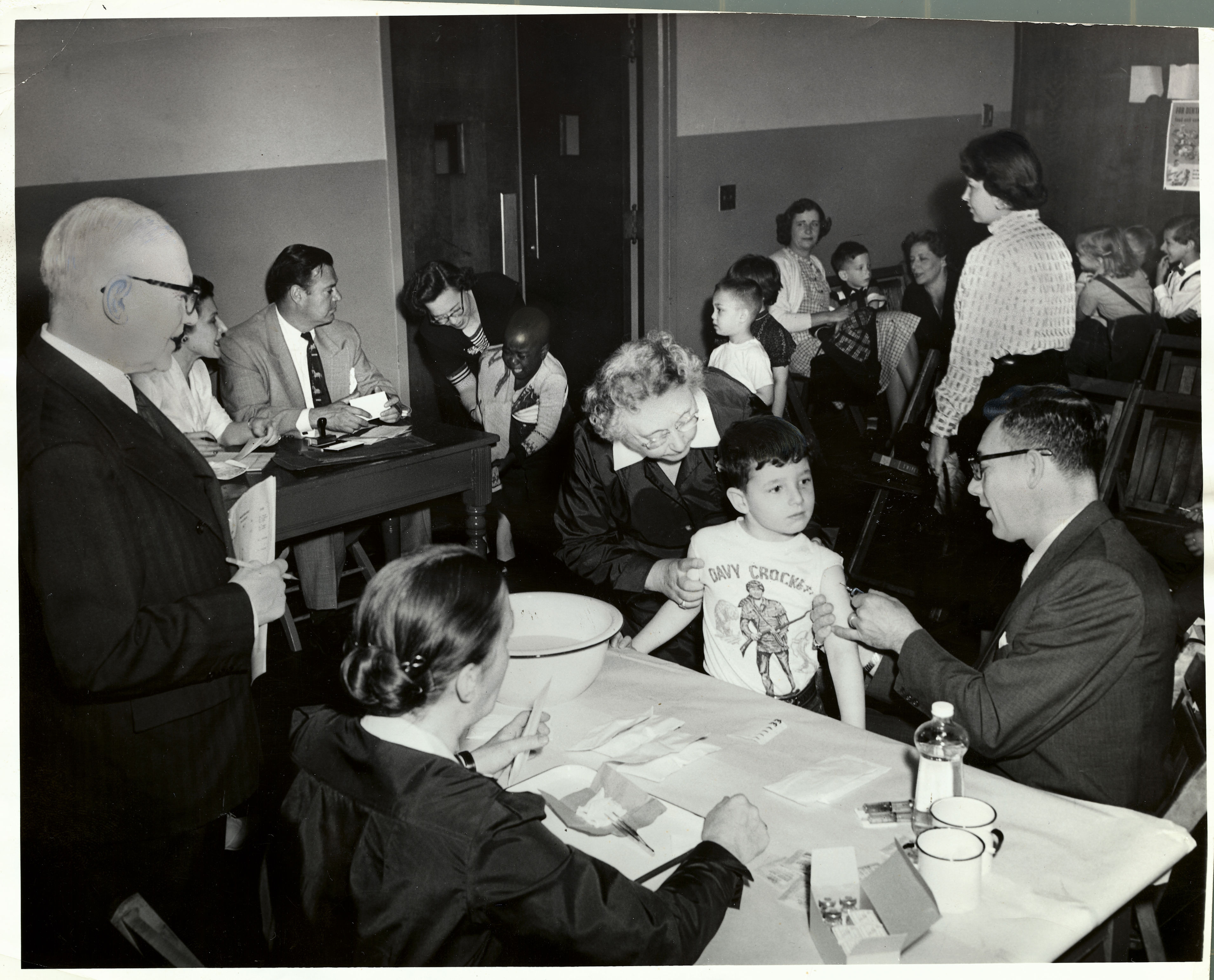 Black and white photo of a child in a "Boy Corcho" shirt getting checked by a man at a medical station, surrounded by adults and other children