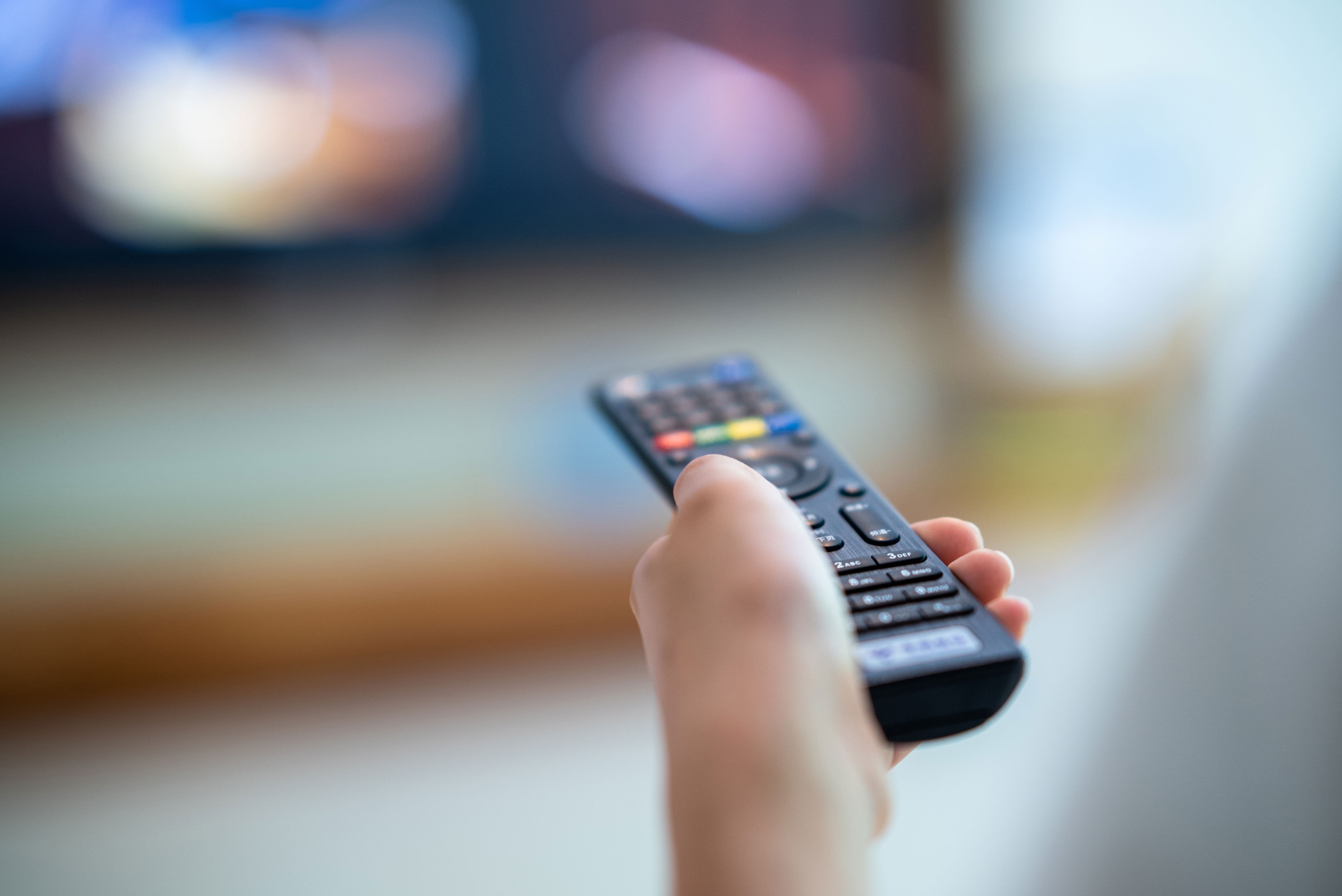 Person holding a remote control, pointing it at a blurred TV screen in the background