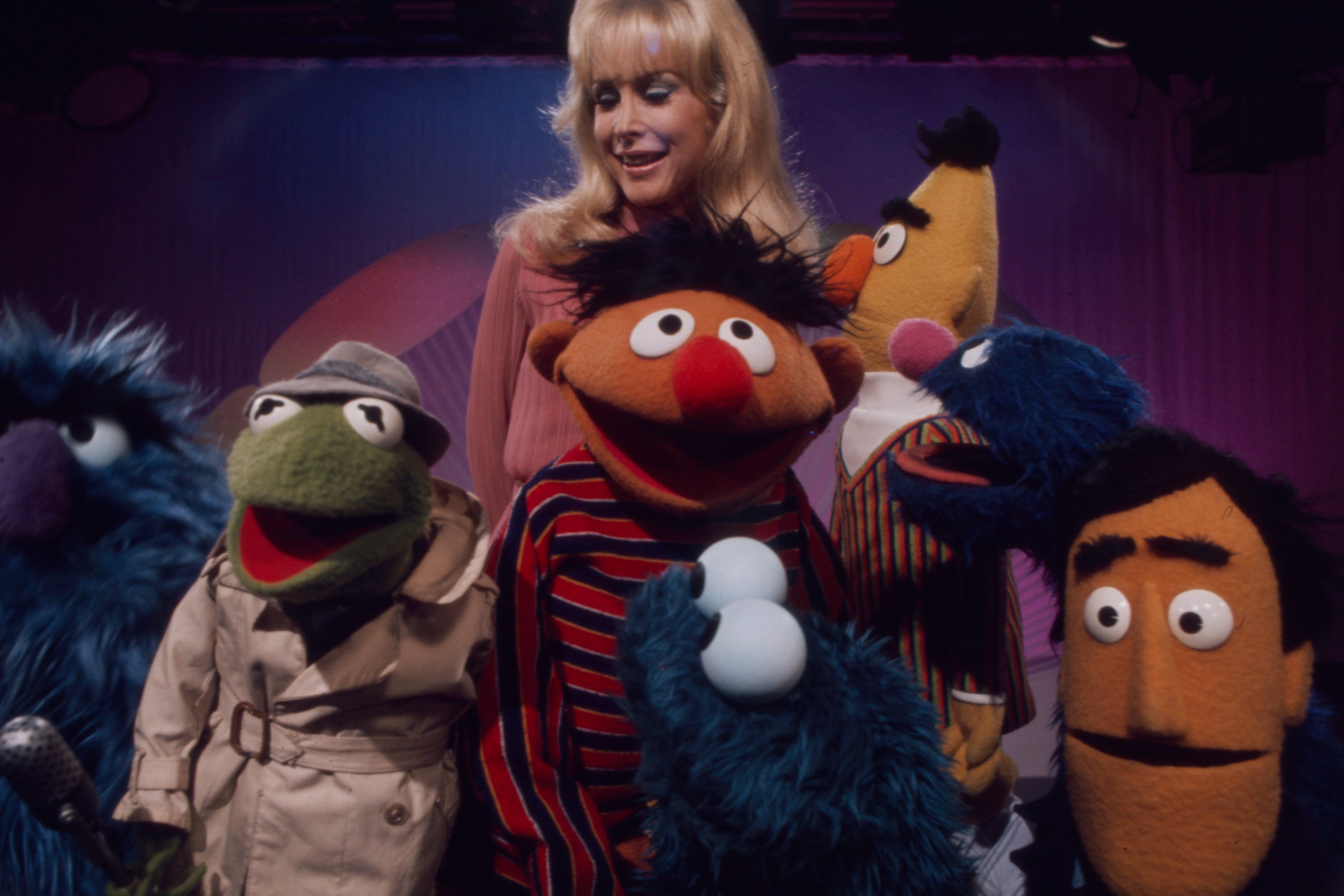 A group of Muppet characters with a woman smiling behind them, including Kermit in a trench coat and Ernie in a striped sweater